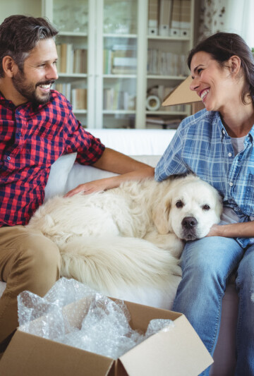 Man and woman sitting on sofa with their large white dog amongst moving boxes
